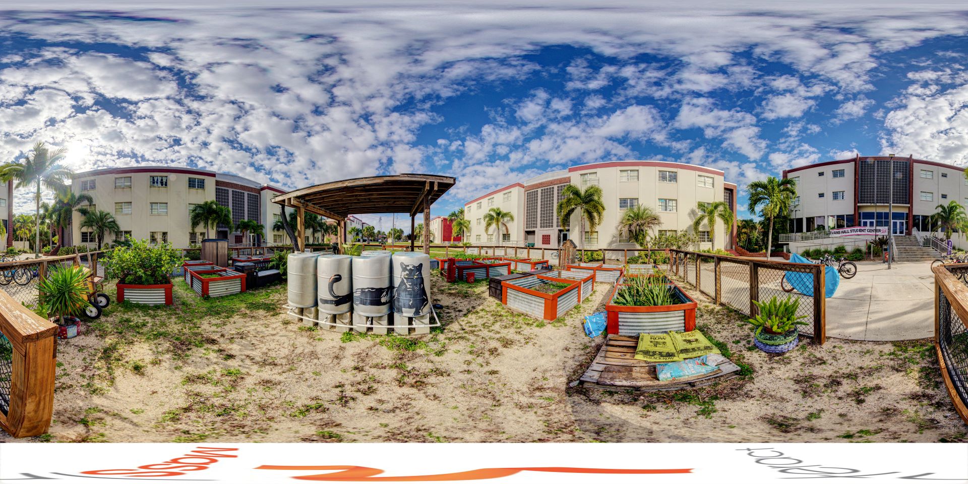 Panoramic view of a college campus outdoor area featuring Ethos Garden with raised plant beds, a covered seating area, and student buildings in the background under a partly cloudy sky